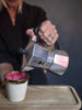 A person pouring coffee from a silver aluminum moka pot into a small ceramic cup with a gold rim and pink interior, set on a wooden tray against a dark background.