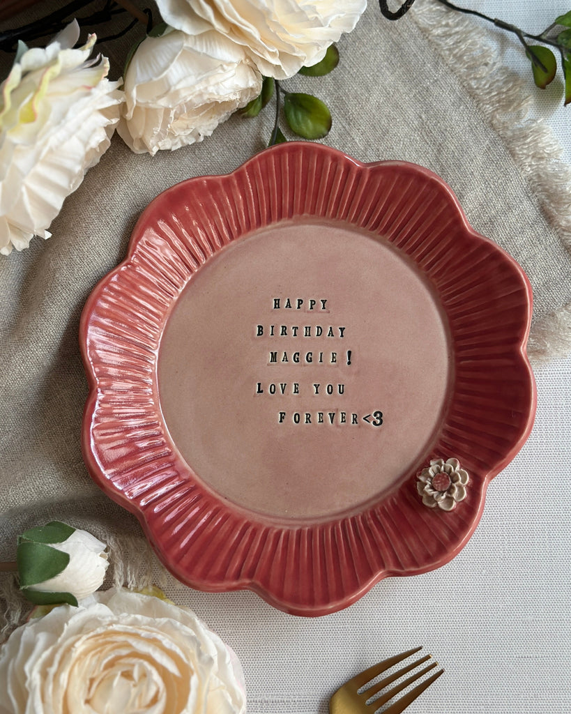 A scalloped ceramic plate in terracotta red with a dusty pink center, featuring the text 'HAPPY BIRTHDAY MADDIE! LOVE YOU FOREVER' and a small floral embellishment. The plate is styled in a flat-lay setting with white roses, a gold fork, and a beige linen runner.