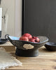 A dark charcoal ceramic bowl with a textured, rustic finish and a small beige decorative accent, filled with red apples on a wooden table. A matching dark bowl sits in the background.