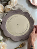 A scalloped ceramic plate with a muted lavender-gray exterior and a warm cream interior, held by a hand. The plate features black printed text reading 'YOU ARE SPECIAL TODAY' and 'BLATLOCK FAMILY CELEBRATION PLATE' surrounding a small diamond icon.