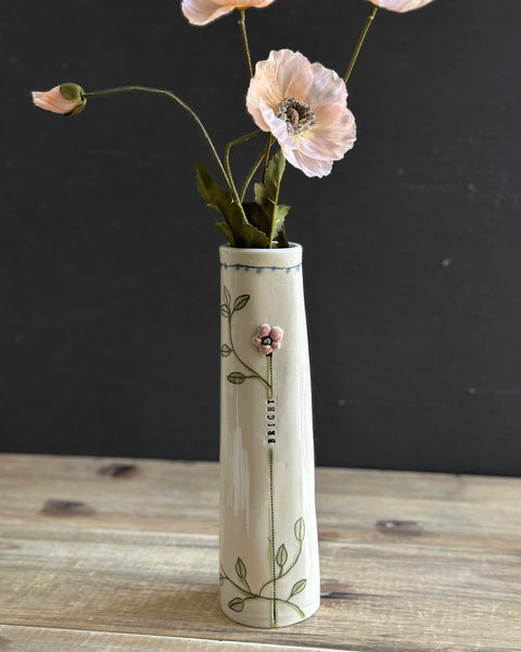 A tall, slender ceramic vase with a cream-colored glaze, decorated with green leafy vines and a small pink flower. It is filled with artificial light pink poppy flowers and is set on a rustic wooden surface against a dark gray background.