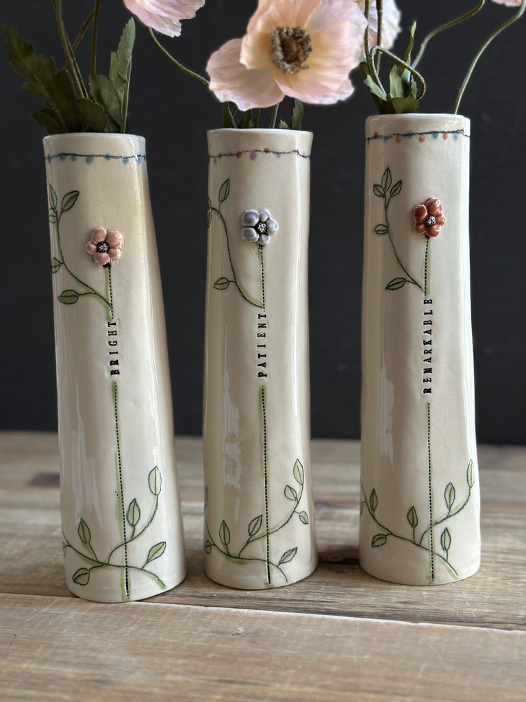 Three tall cream ceramic vases with green vine and flower illustrations, displayed on a wooden surface against a dark background with pink flowers in the vases.