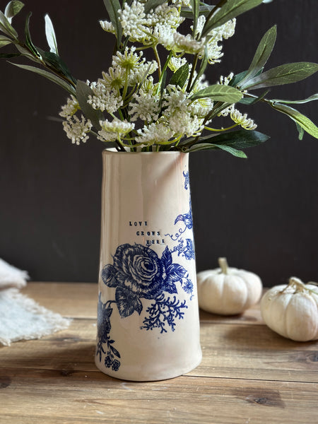 A tall cream ceramic vase with blue floral and rose illustrations, filled with white flowers and green leaves, set on a rustic wooden table with small pumpkins in the background.