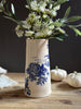 A tall cream ceramic vase with blue floral and rose illustrations, filled with white flowers and green leaves, set on a rustic wooden table with small pumpkins in the background.