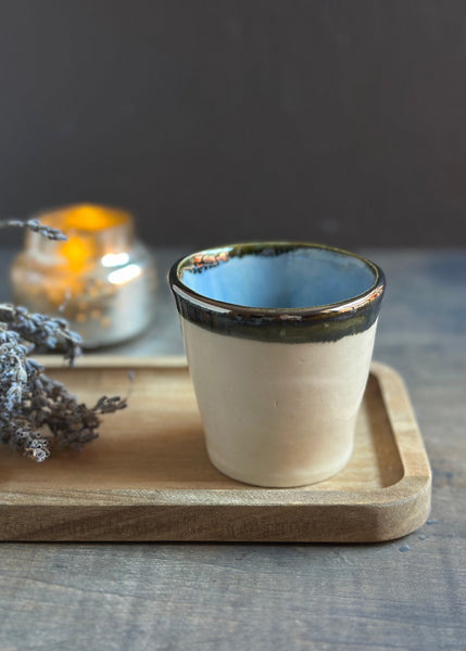 A handmade ceramic cup with a cream-colored exterior, a dark green glazed rim, and a light blue interior, displayed on a wooden tray with dried lavender and a blurred candle in the background.