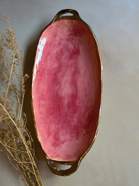 An oval ceramic serving platter with a glossy pink marbled glaze and metallic gold handles, styled with dried botanical stems on a neutral gray background.