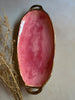 An oval ceramic serving platter with a glossy pink marbled glaze and metallic gold handles, styled with dried botanical stems on a neutral gray background.