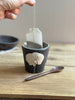 Tea bag being placed into a black ceramic cup with a decorative handle on a wooden surface.