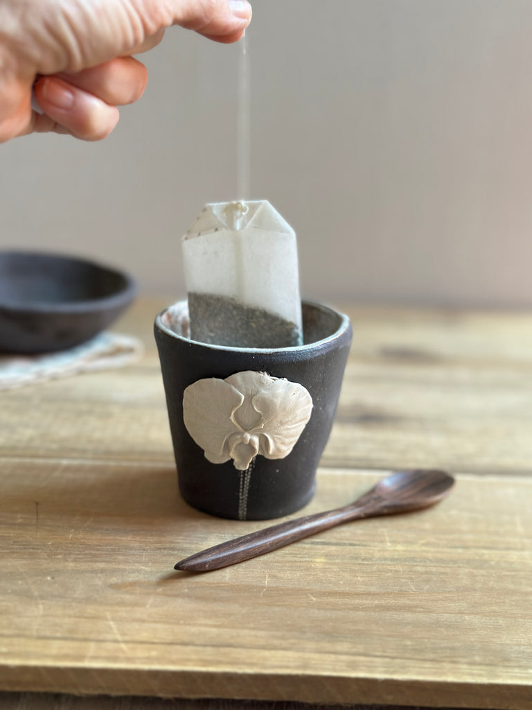 Tea bag being placed into a black ceramic cup with a decorative handle on a wooden surface.