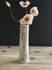 A tall, narrow cream-colored ceramic vase with green floral illustrations and the brand name 'PARIST' printed vertically, holding four pale pink poppy flowers against a dark background and wooden tabletop.