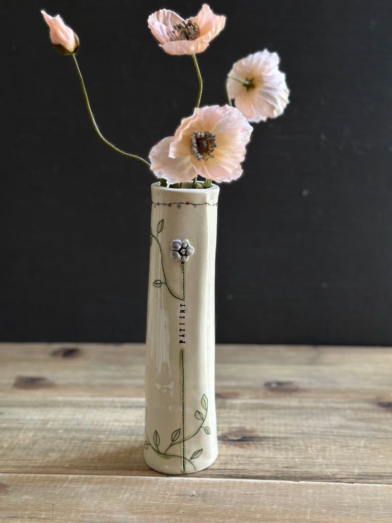 A tall, narrow cream-colored ceramic vase with green floral illustrations and the brand name 'PARIST' printed vertically, holding four pale pink poppy flowers against a dark background and wooden tabletop.
