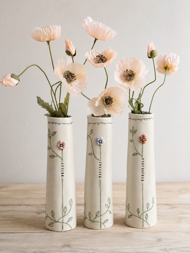 Three tall cream ceramic vases with delicate floral illustrations, arranged on a light wood surface against a neutral light gray wall, holding pale pink poppy flowers.