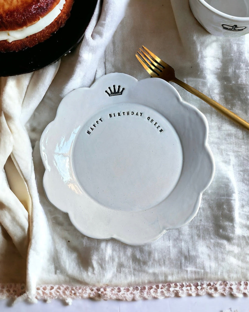 White decorative plate with text on a white tablecloth with a cake and bowl in the background.