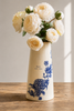 A cream-colored ceramic vase with blue floral illustrations and the text 'GAL EST. 1994', filled with cream-colored roses, set on a rustic wooden table against a neutral beige wall.